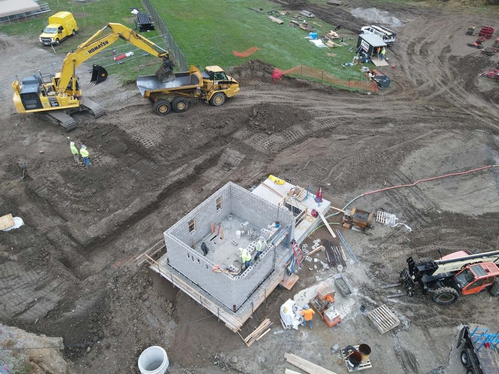 Aerial view of an active construction site showing an excavator loading soil into a dump truck and workers building a small concrete block structure in the foreground.