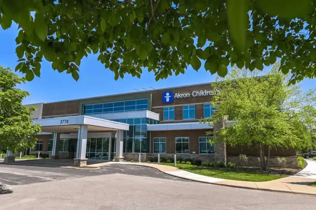 Exterior view of an Akron Children’s medical building with a glass entrance canopy and landscaped trees under a clear blue sky.