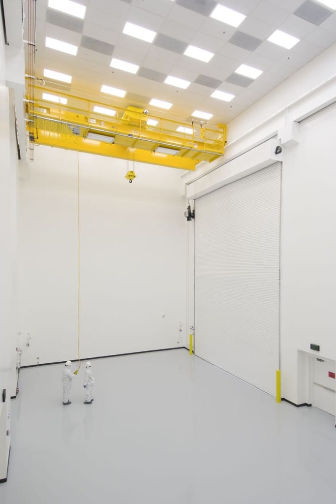 Two technicians in full cleanroom suits standing beneath a bright yellow overhead crane inside a white industrial testing chamber.