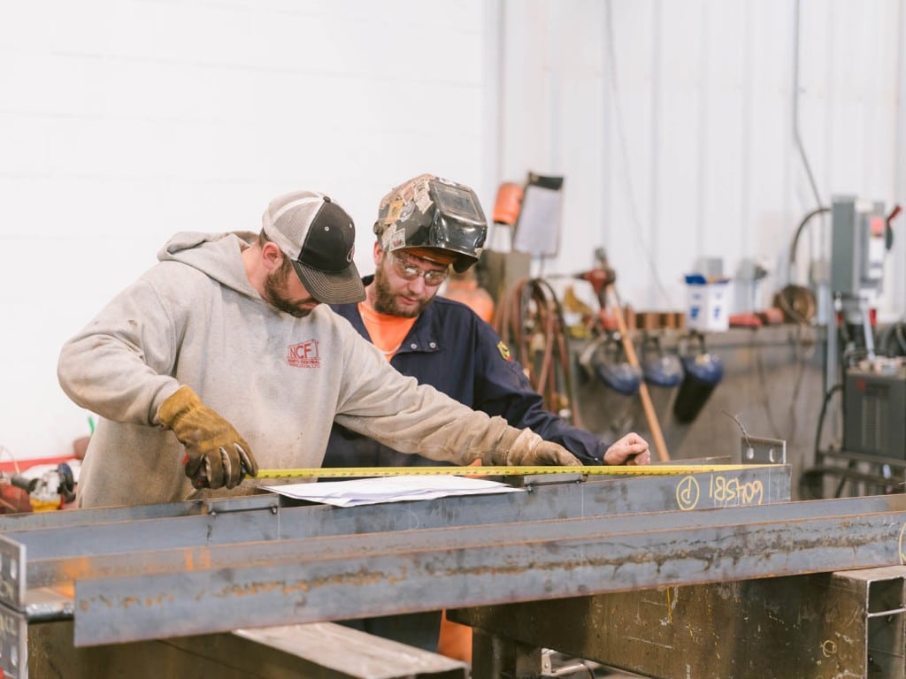 Two workers measuring a steel beam in a fabrication shop.
