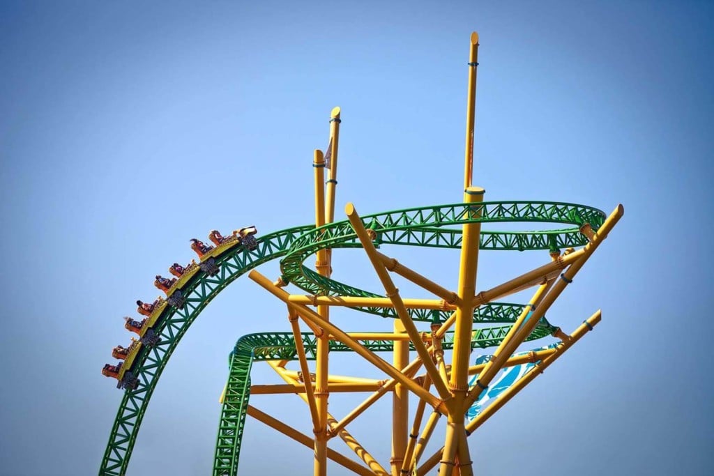 Roller coaster train cresting a steep green track supported by yellow steel columns against a clear blue sky.