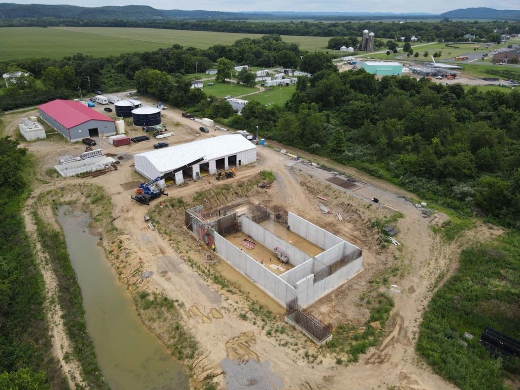 Aerial view of a rural construction site with a concrete foundation, a white metal building, and surrounding equipment.