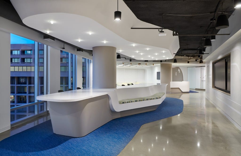 “Modern office reception area featuring a long sculpted white reception desk with built-in curved seating, blue carpet pathway, floor-to-ceiling windows, and a minimalist open-ceiling design with recessed and pendant lighting.