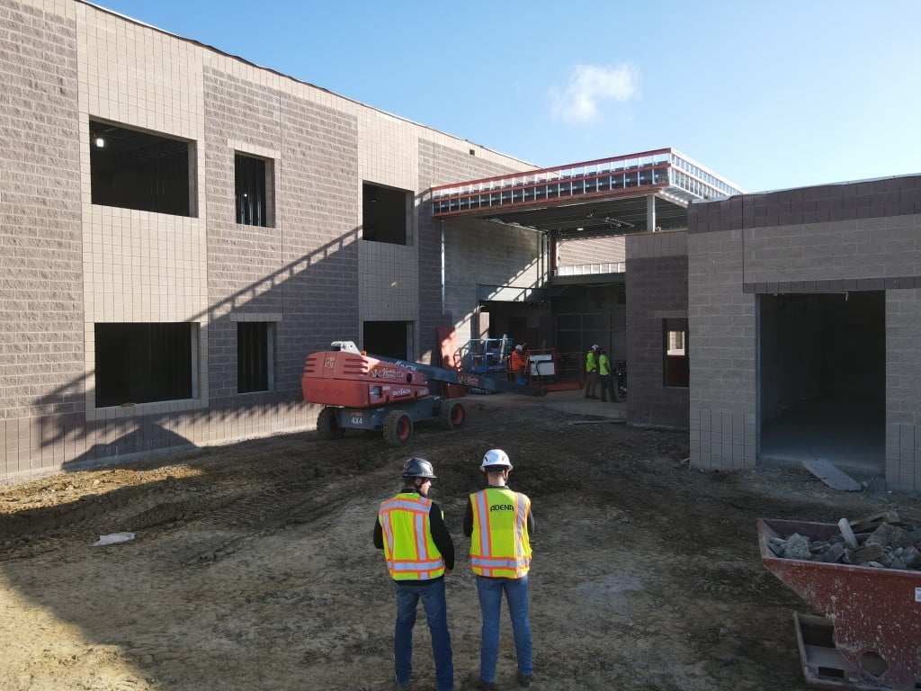 Two construction workers in high-visibility vests and hard hats standing in front of a masonry school building under construction, with a lift and other workers visible in the background.