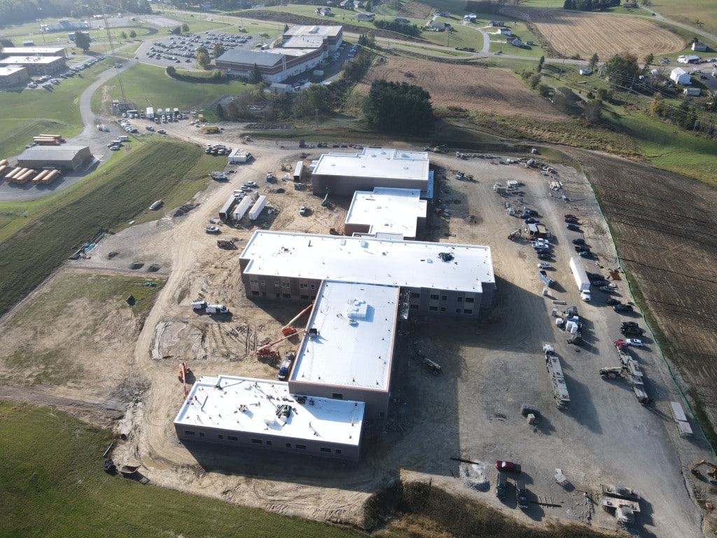 Aerial view of a large school building under construction, with multiple connected wings, construction vehicles, and surrounding farmland.