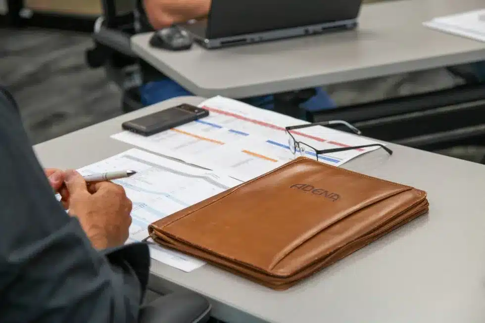 Man with papers and leather folder on desk that says Adena