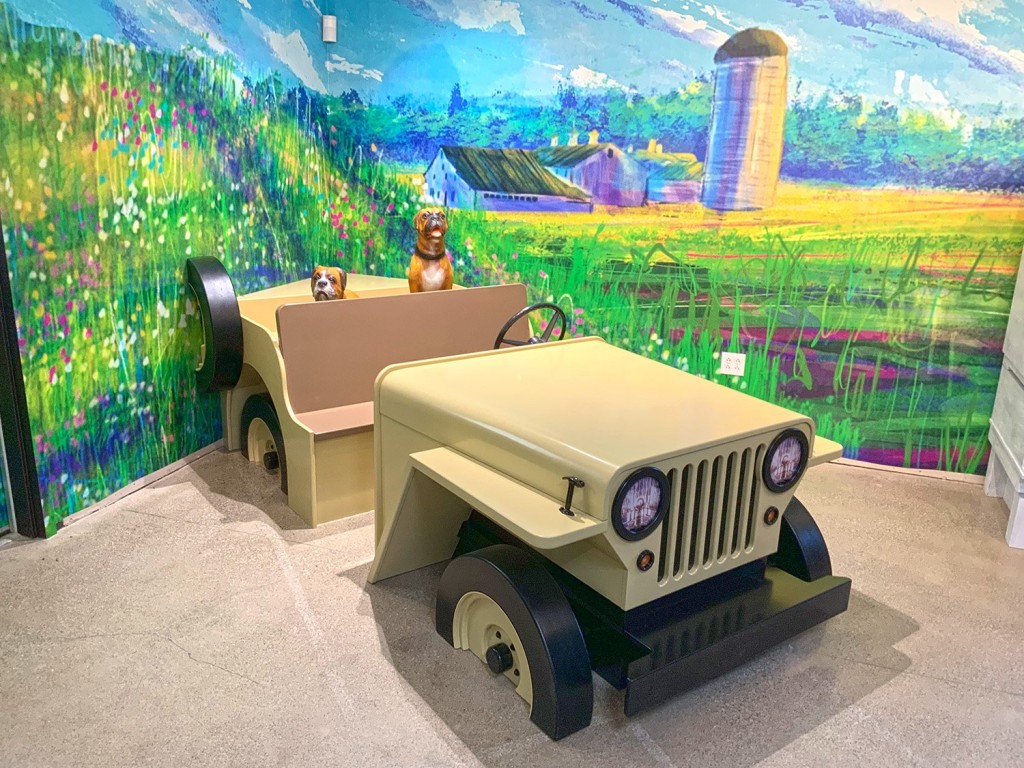 Interactive exhibit with two sculpture dogs sitting in car made of wood and a mural of a field with flowers and grass
