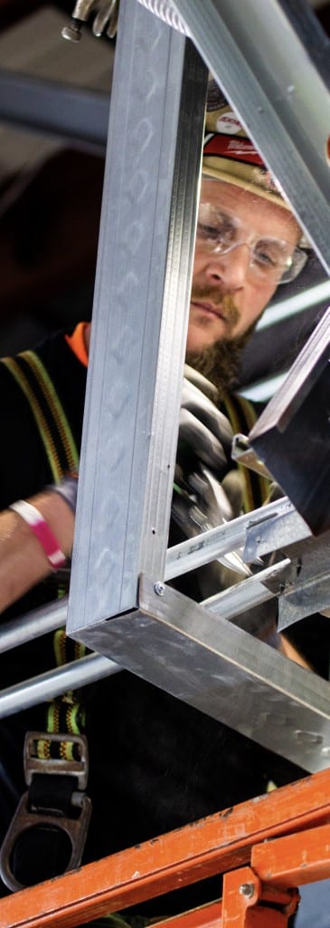 Construction worker wearing safety glasses and a hard hat fastening a metal framing connection while secured with a harness.