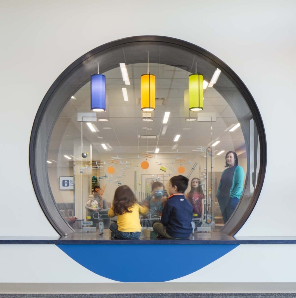 Children sitting on the floor inside a glass-walled room viewed through a large circular window, with an adult standing nearby and three colorful hanging lights above.