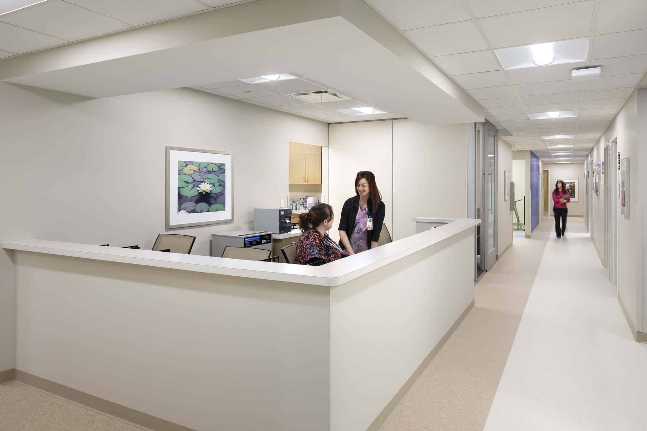 Medical office reception desk with two staff members speaking behind a curved counter and a hallway extending to the right.