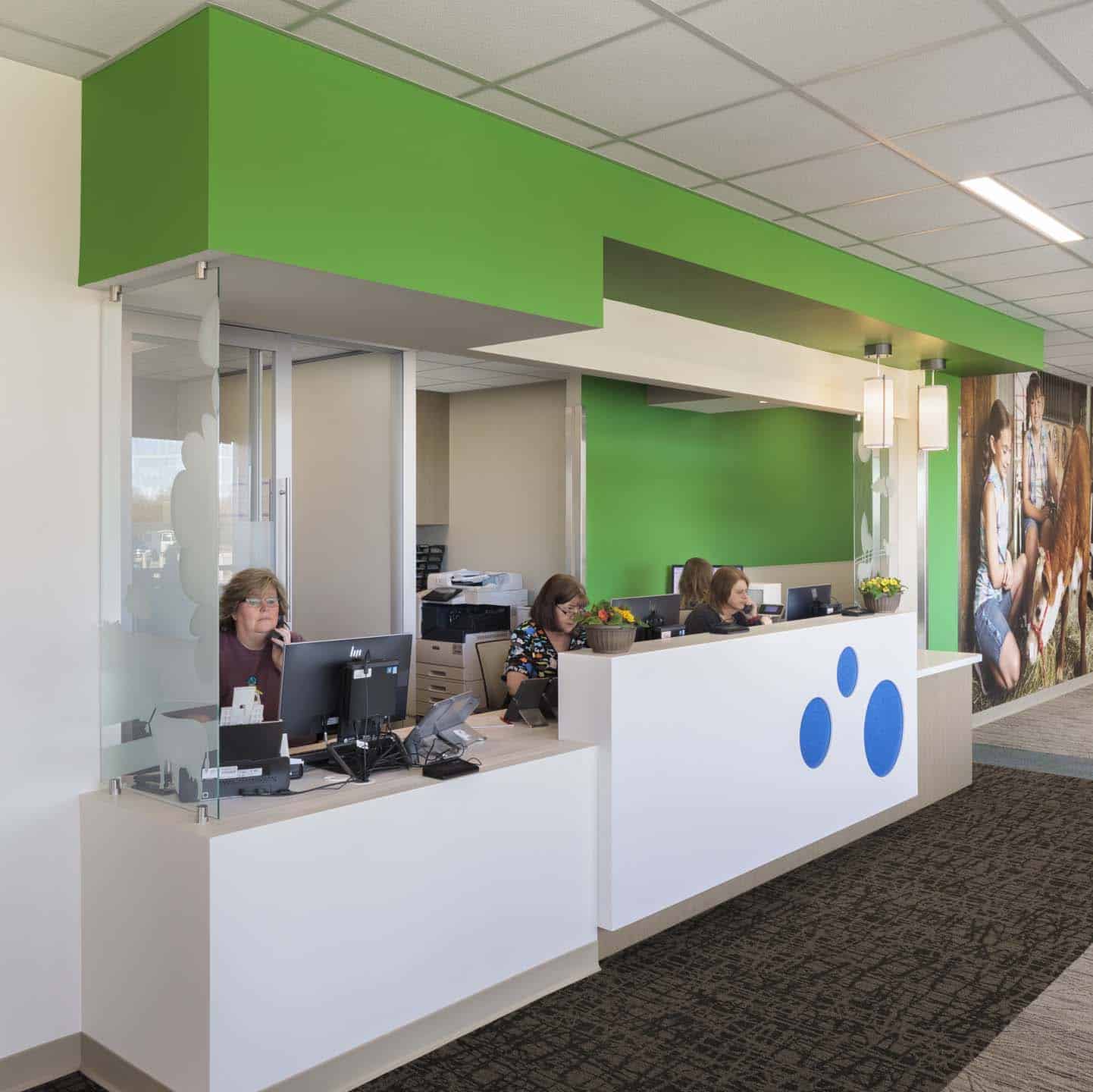 “Front desk reception area with three staff members behind a white counter with blue dot graphics and a green accent wall.