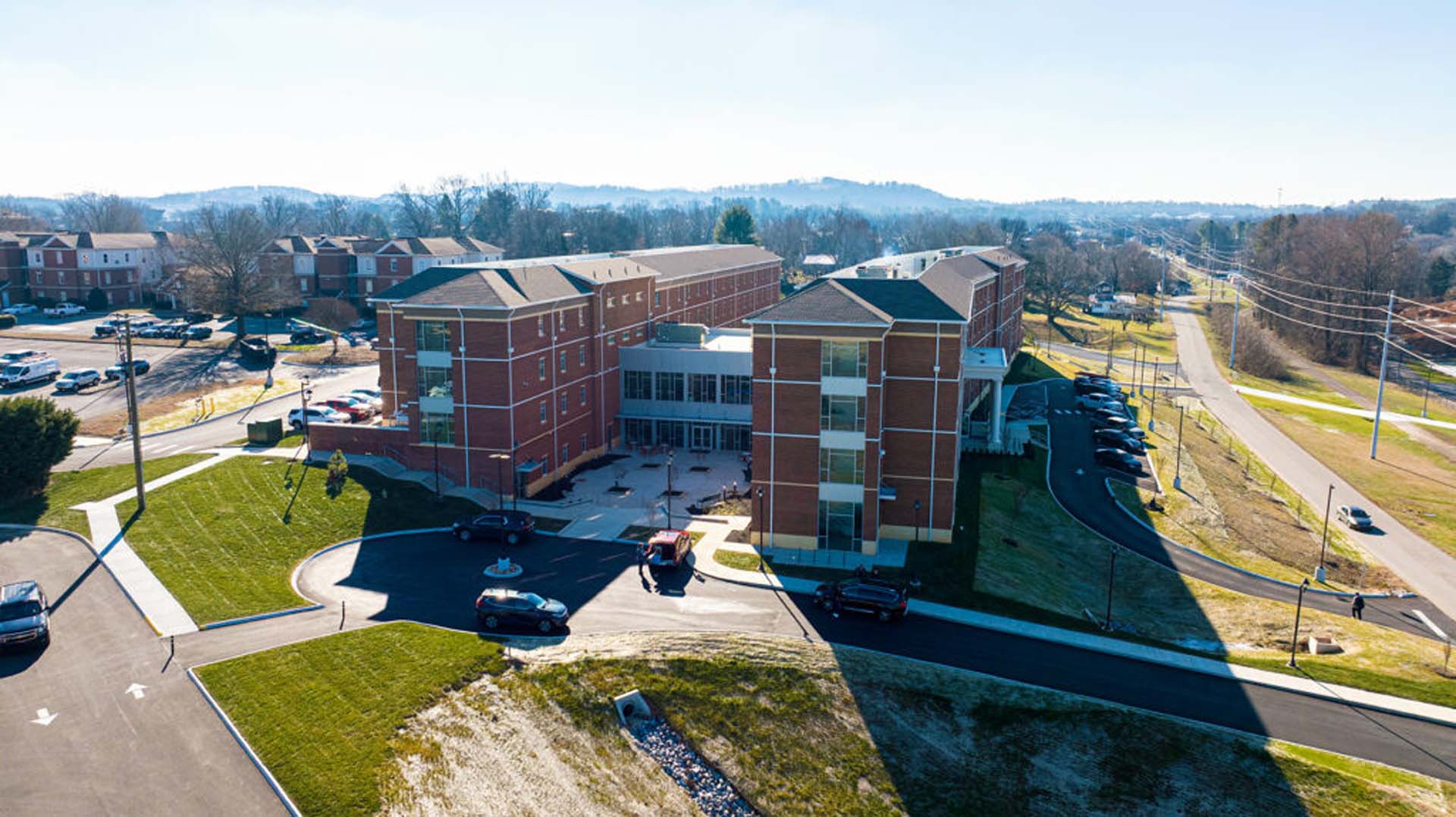 Aerial view of a multi-story brick residential complex with a central courtyard and surrounding parking, set along a curved roadway in a suburban landscape.
