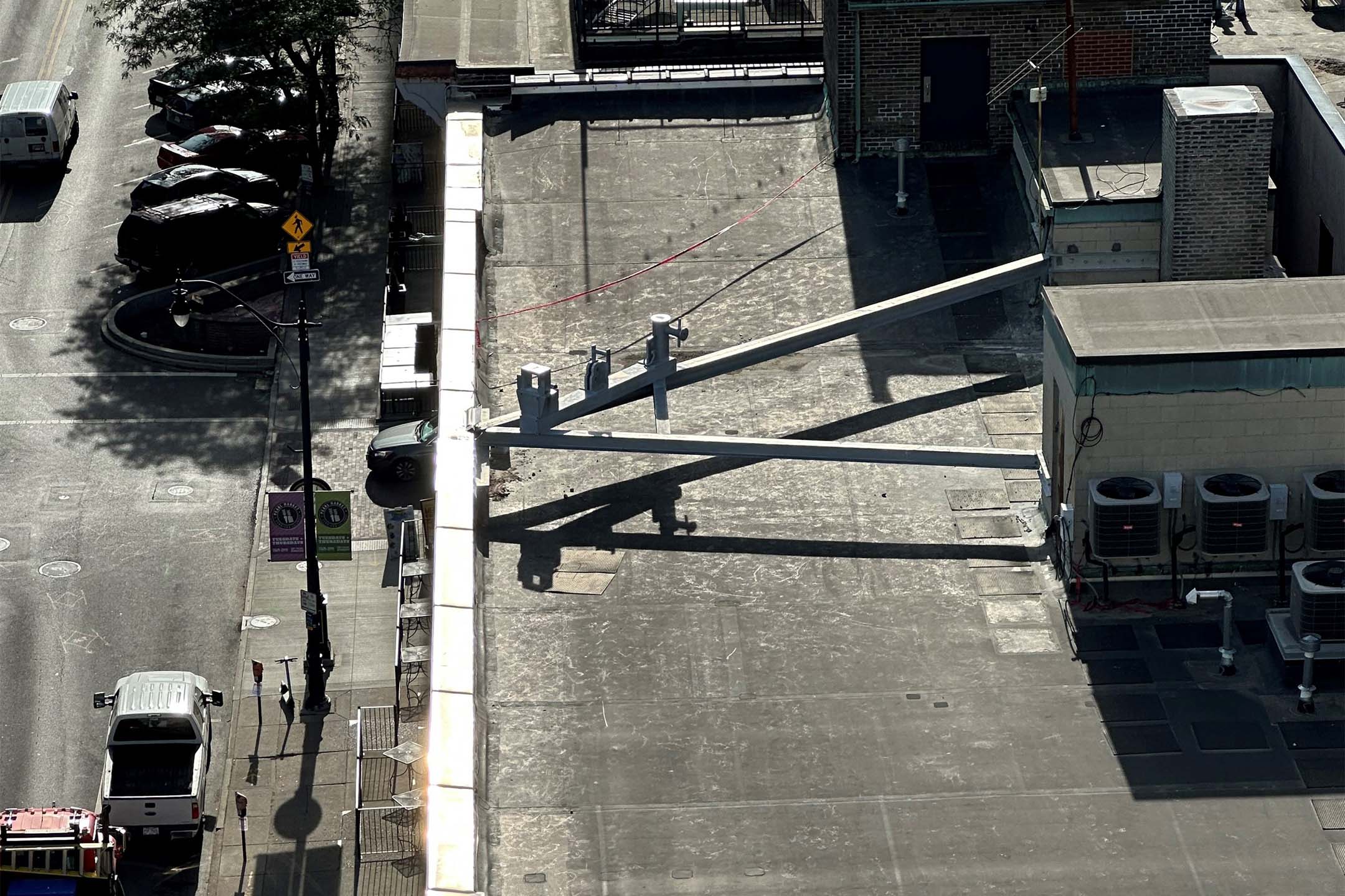 Aerial view of a rooftop and adjacent alley showing a large metal beam structure extending across the roof, casting long shadows, with HVAC units and a parked van nearby.