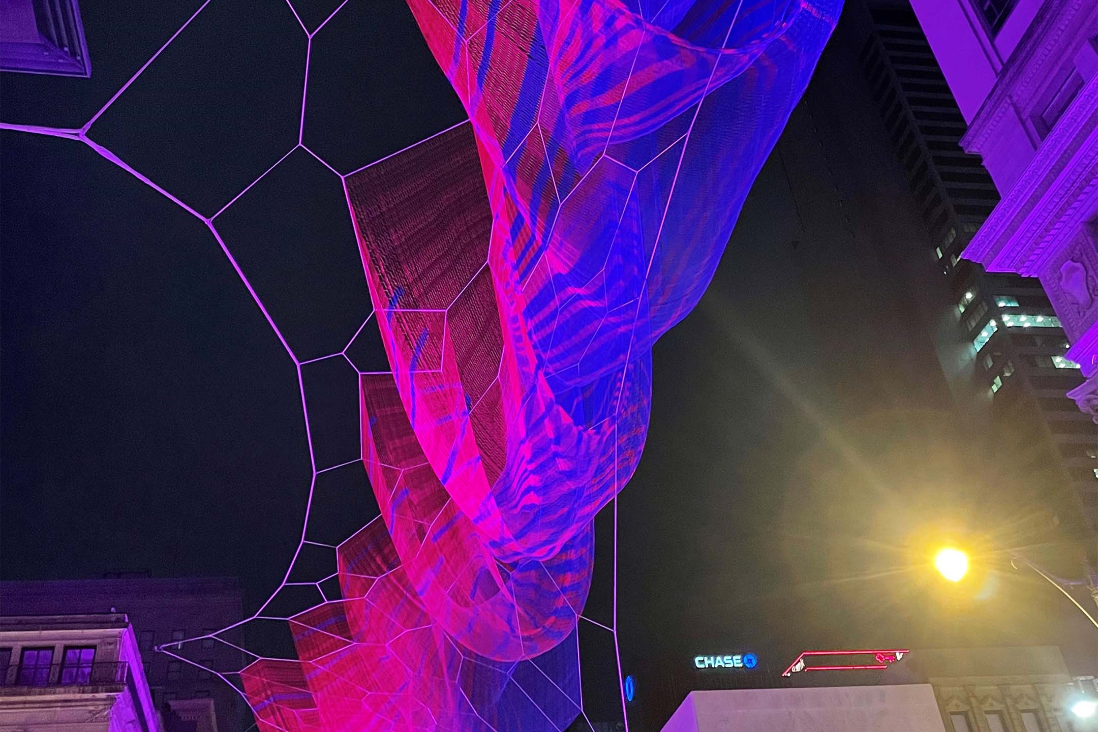 Nighttime view of a large, glowing pink and purple mesh-like light installation stretched across city buildings against a dark sky.