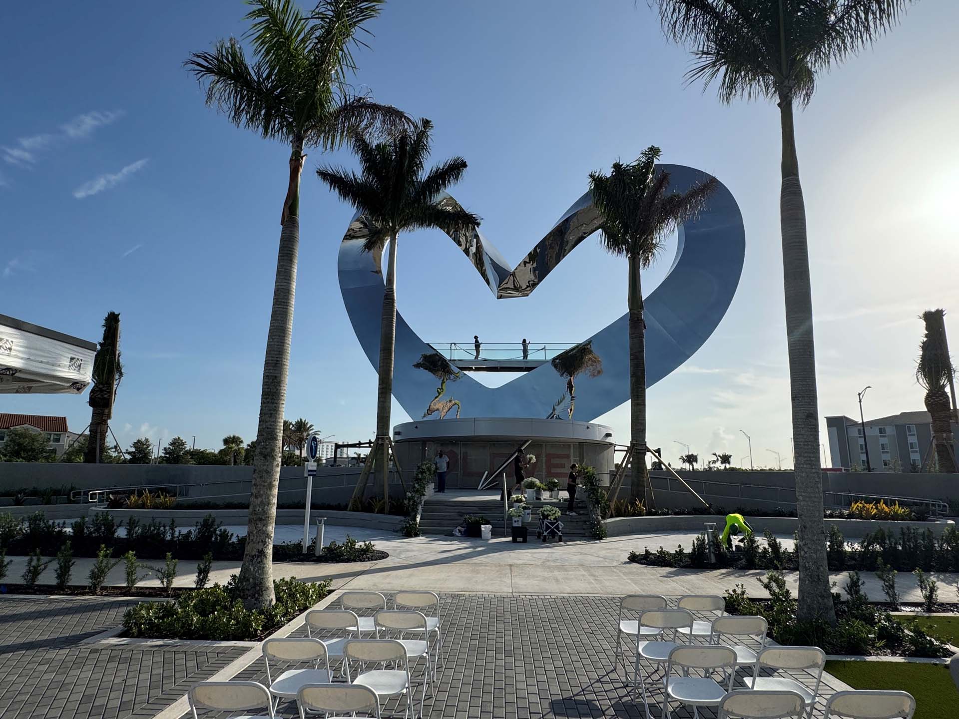 Curved reflective sculpture in a small plaza framed by palm trees.