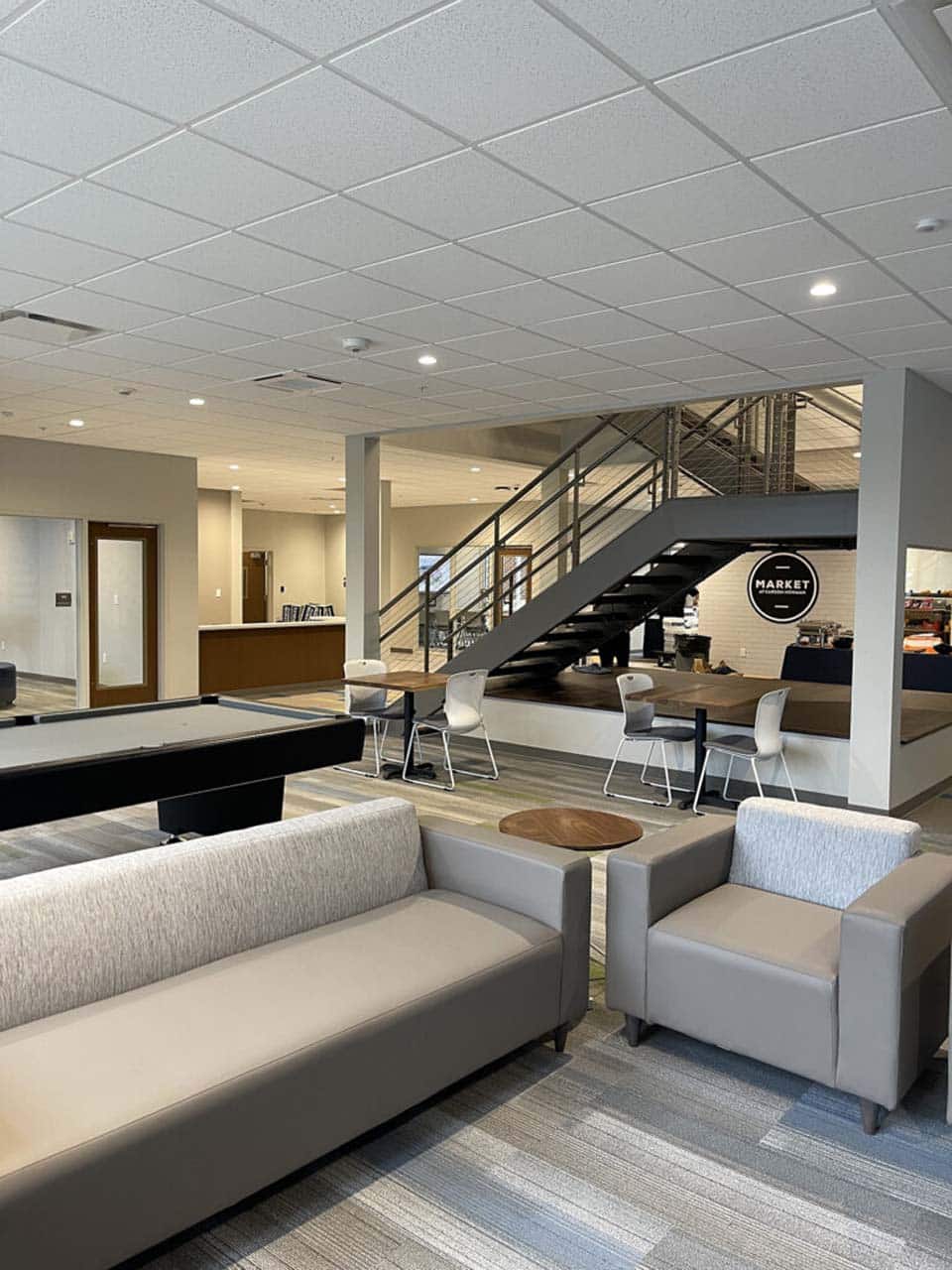 Modern office lounge with seating, a pool table, and a staircase leading to a café area.