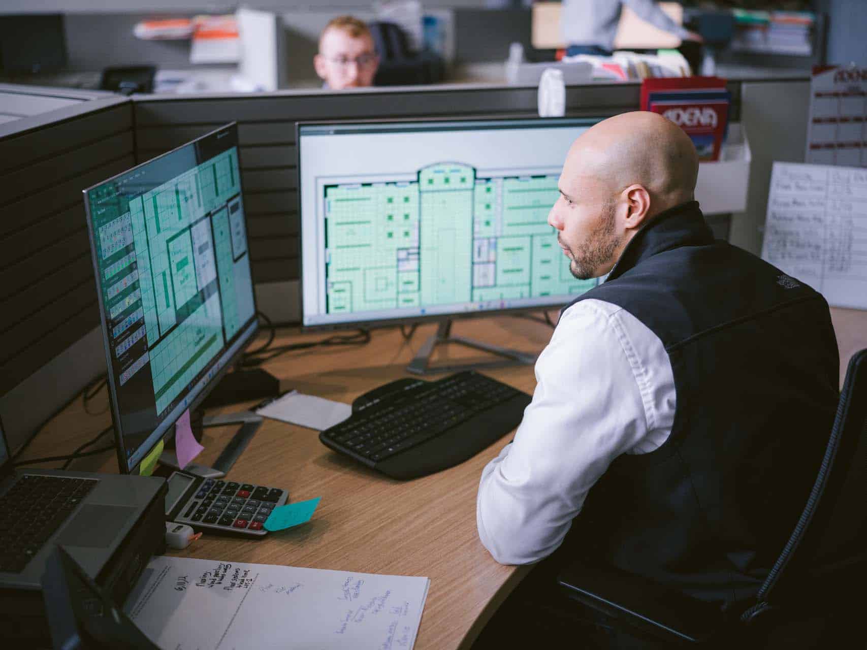 Employee reviewing building floor plans on dual computer monitors at a desk.